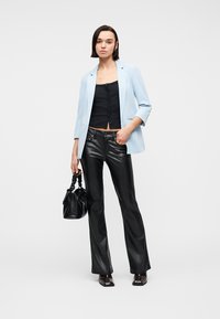 Γυναίκα που στέκεται, φορώντας ένα ανοιχτό μπλε σακάκι, μαύρο μπλουζάκι με κουμπιά, μαύρο δερμάτινο παντελόνι, κρατώντας μια μαύρη τσάντα, με καφέ σκούρα παπούτσια.