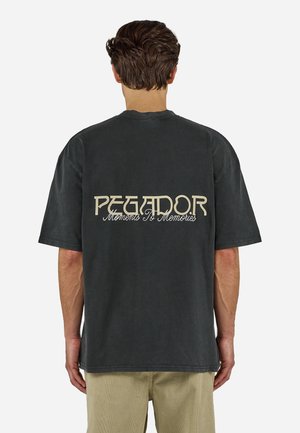 Άνδρας που φοράει μαύρο φαρδύ T-shirt με το κείμενο "PEGADOR Moments To Memories" στην πλάτη, σε συνδυασμό με ανοιχτό μπεζ παντελόνι.