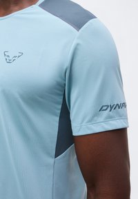 Ανοιχτό μπλε αθλητικό κοντομάνικο με σκούρα γκρι πάνελ στους ώμους και τις πλευρές, ένα στυλιζαρισμένο λογότυπο ζώου στο στήθος και τη λέξη "DYNA" στο μανίκι.