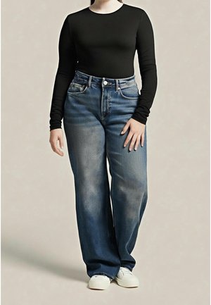 Person trägt ausgewaschene, weit geschnittene blaue Jeans, ein schwarzes, eng anliegendes Langarmshirt und weiße Turnschuhe und steht vor einem schlichten Hintergrund.