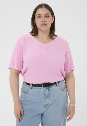 Woman wearing a pink V-neck short-sleeve top tucked into light blue jeans with a black belt, silver earrings, ring, and bracelet.