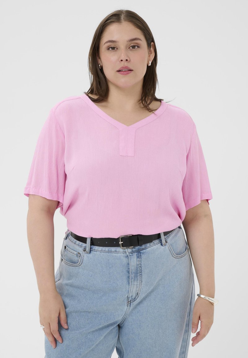 Femme portant un haut rose à manches courtes en V, rentré dans un jean bleu clair avec une ceinture noire, des boucles d'oreilles argentées, une bague et un bracelet.