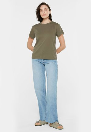 Olivengrünes Baumwoll-T-Shirt mit kurzen Ärmeln und rundem Halsausschnitt, kombiniert mit hellblauen, weit geschnittenen Jeans und flachen Sandalen, die minimale Designdetails aufweisen.