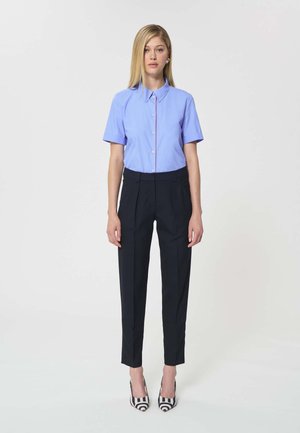 Femme debout portant une chemise à manches courtes bleu clair boutonnée, un pantalon noir sur mesure et des talons hauts rayés noir et blanc.