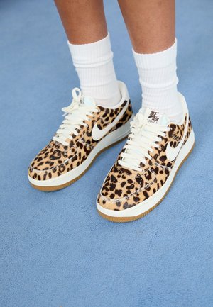 Persoon met witte sokken en Nike Air Force 1 sneakers met luipaardprint, staand op een blauwe tapijtvloer.