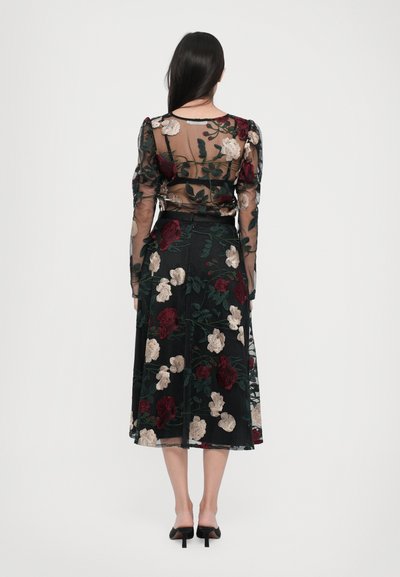 Doorzichtig zwart jurk met lange, puffed mouwen; heeft meerkleurige bloemenborduursels in rood, crème en groen; maxi-lengte en uitlopende silhouet.