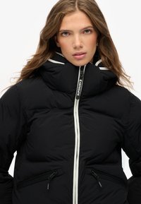 Chaqueta acolchada negra con un cuello alto, cremallera blanca y dos bolsillos frontales con cremallera. Presenta una textura suave y detalles de contraste elegantes.