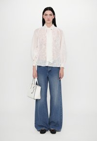 Blusa bianca trasparente con ricami floreali, abbinata a jeans a gamba larga blu, mentre tiene una piccola borsa bianca e indossa ballerine nere.