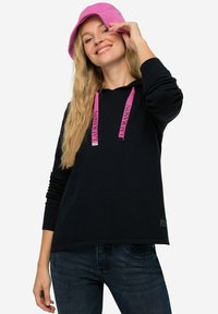 Felpa nera con cordini rosa, caratterizzata da un logo, maniche lunghe e una vestibilità rilassata. Indossata insieme a un cappello a secchiello rosa e pantaloni in denim.
