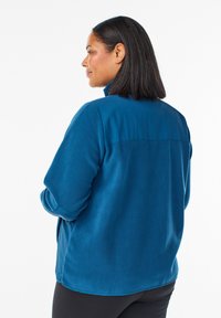 Kvinna med axellångt svart hår iförd en blå fleecejacka och svarta byxor, vänd åt sidan med händerna i fickorna.