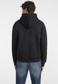 Sort hettegenser laget av myk stoff, med en stor hette og ribbestrikkede mansjetter. Sett fra baksiden, kombinert med lyseblå jeans.