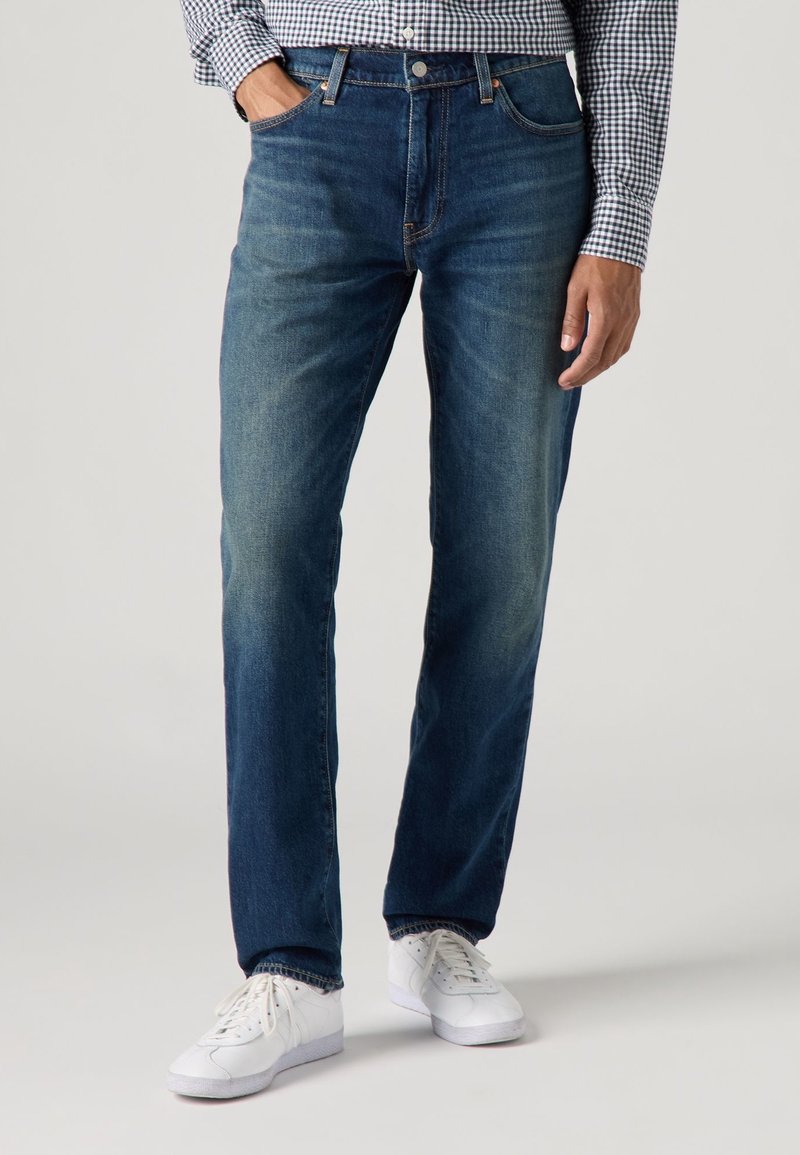Homme portant un jean bleu foncé, des baskets blanches et une chemise à carreaux noir et blanc, une main dans la poche.