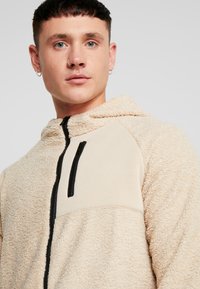 Beige fleece hættetrøje med et kontrasterende glat stofpanel, lynlås foran og sort lynlåsdetalje på brystlommen. Tekstureret finish.
