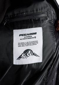 Svart jakkeinnside som viser et hvitt Pegador yttertøymerke med fjellgrafikk og beskrivende tekst om produktet som er eventyrlystent.