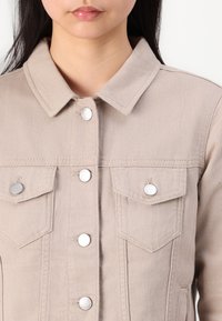 Beige denim jakk, millel on krae, esi nupuga sulgemine ja kaks rinnataskut, kus on hõbedased metallnupud. Tekstureeritud kangas.
