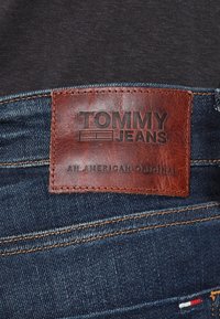 Jeans i denim har en brun läderlapp med präglad "TOMMY JEANS" och "AN AMERICAN ORIGINAL", placerad mot en mörkblå tyg med orange sömmar.