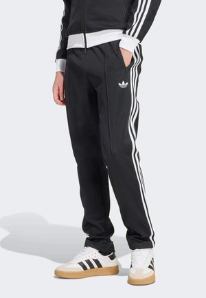 Person trägt schwarze Adidas Trainingshose mit weißen Seitenstreifen und Logo, kombiniert mit weißen Adidas Sneakern mit schwarzen Streifen.