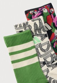 Trzy złożone skarpetki z prążkowanymi mankietami: zielono-białe paski, kremowe z czarnym abstrakcyjnym napisem oraz wielokolorowe papugi na czarnym tle.