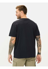 Zwarte katoenen t-shirt met korte mouwen, een ronde halslijn en een ontspannen pasvorm. Heeft zichtbare tatoeages op de rechterarm.