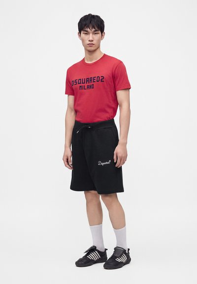 Jeune homme portant un t-shirt rouge Dsquared2 Milano, un short noir, des chaussettes blanches et des baskets noires Adidas, debout devant un fond uni.