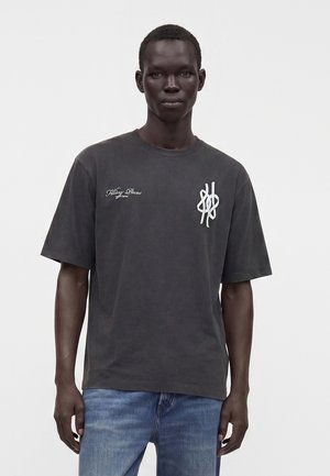 Ung mand iført en mørkegrå oversize T-shirt med hvid tekst og logo på brystet, kombineret med blå jeans, stående mod en ensfarvet baggrund.