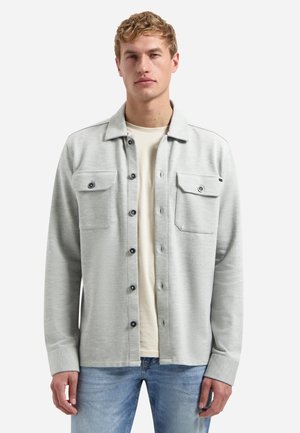 Blonder Mann trägt ein hellgraues Hemdjacke mit Brusttaschen über einem beigefarbenen T-Shirt und blauen Jeans, steht vor weißem Hintergrund.