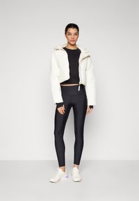 Vit kortärmad pufferjacka över en svart långärmad topp, matchad med svarta leggings och vita sneakers med lila detaljer. Släta texturer.