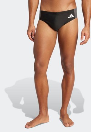 Male model wearing black Adidas swim briefs standing barefoot against a plain light background.