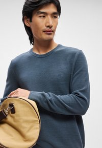 Blå teksturert strikkegenser med rund hals, båret av en person som holder en rund, beige veske med brun stropp og detaljer.