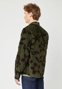 Camicia in corduroy verde con motivi floreali neri, maniche lunghe e polsini abbottonati. La vista posteriore mette in risalto una vestibilità rilassata e un tessuto testurizzato.