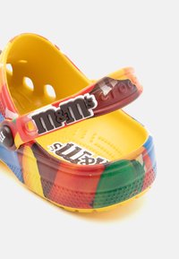 Des Crocs multicolores avec un design en plastique brillant, un intérieur jaune, une sangle ajustable et le logo M&M's sur la bande.