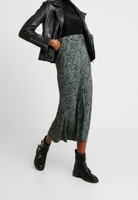 Grüne Leopardenmuster-Midi-Rock, fließendes Design, kombiniert mit einem schwarzen gerippten Oberteil und einer schwarzen Lederjacke. Schwarze Ankle-Boots mit Akzenten.