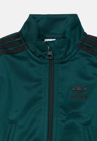 Tirkizna adidas jakna za trening s visokom ovratnikom, crnim akcentima, zatvaranjem na patentni zatvarač i tri crne pruge na ramenima.