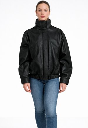 Femme debout, tournée vers l’avant, vêtue d’une veste en cuir noire à col haut et d’un jean bleu, sur fond blanc.