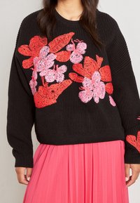 Svart stickad tröja med röd och rosa blommönsterbroderi, kort design med långa ärmar, matchad med en plisserad rosa kjol.