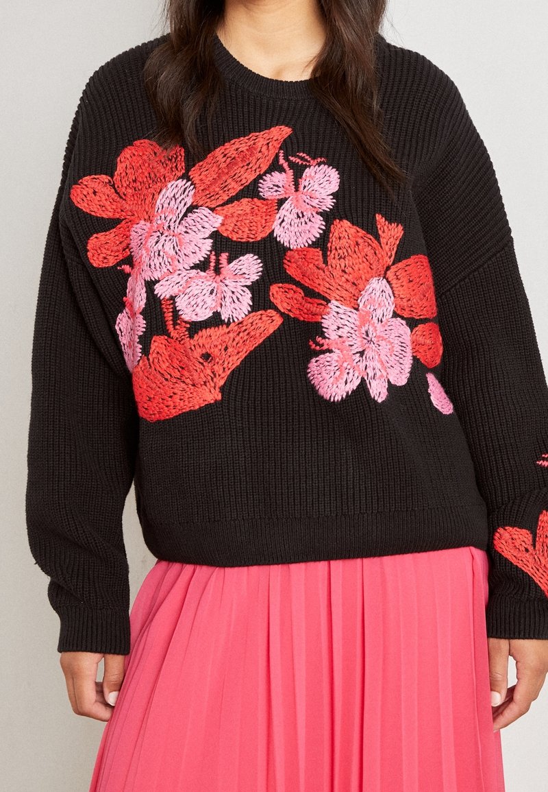 Svart stickad tröja med röd och rosa blommönsterbroderi, kort design med långa ärmar, matchad med en plisserad rosa kjol.