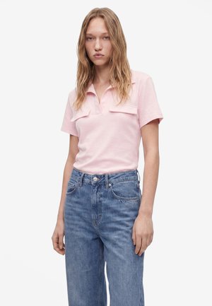 Jeune femme aux longs cheveux blonds portant un t-shirt rose clair à manches courtes et un jean bleu taille haute sur un fond uni.