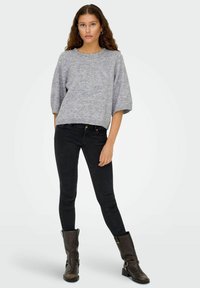 Grijze gebreide sweater met wijde mouwen, ronde halslijn en decoratieve afwerking. Gecombineerd met zwarte slim-fit jeans en donkerbruine enkellaarzen.