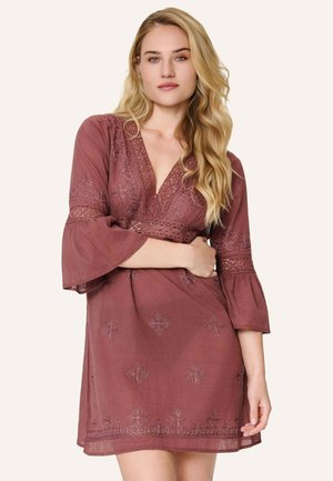 Femme aux longs cheveux blonds portant une robe mauve à col en V avec des détails en dentelle et des manches trois-quarts évasées, debout devant un fond clair.