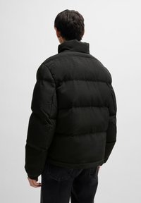 Schwarze Winterjacke mit hohem Kragen, gestepptem Design und elastischen Bündchen, kombiniert mit dunklen Hosen, von hinten betrachtet.