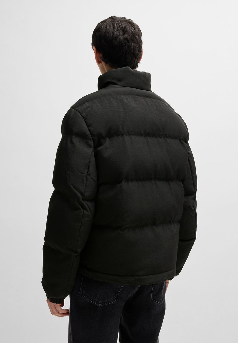 Veste en duvet noire avec un col montant, design matelassé et poignets élastiques, associée à un pantalon sombre, vue de dos.