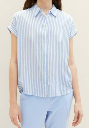 Femme portant une chemise à manches courtes boutonnée à rayures verticales bleu clair et blanc avec un pantalon bleu clair, debout devant un fond uni.