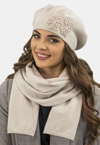 Beret tricotat de culoare bej deschis, decorat cu două accente florale și un fular asortat purtat la gât, având o textură moale și un model discret.