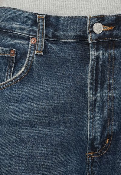 Σκούρο μπλε τζιν με ανθεκτική μέση, χαλκομανίες, πορτοκαλί ραφές και κλασικό σχέδιο με πέντε τσέπες.