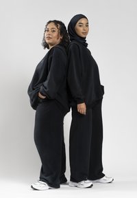 Zwei Frauen stehen Rücken an Rücken, tragen schwarze legere Outfits und weiße Sneaker. Eine mit lockigem Haar, die andere mit einem schwarzen Hijab.