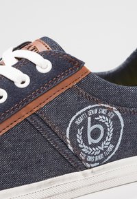 Džínové plátěné tenisky v tmavě modré barvě, s bílými tkaničkami a hnědými koženými doplňky. Vyšitá značka v bílé na vnější straně. Gumová podrážka.