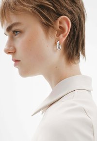 Pendiente de aro de plata con un diseño suave y redondeado. Modelo vistiendo una camisa de color claro con cuello. Pelo corto y texturizado visible.