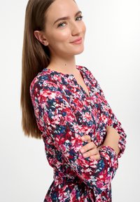 Kvetinové šaty s červenými, ružovými a modrými vzormi. Majú okrúhly výstrih a naťahovacie dlhé rukávy. Hladká textúra s stiahnutým pásom.