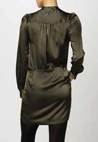 Abito in satin verde oliva con maniche lunghe, dettaglio arricciato sul retro, elastico in vita e tasche laterali. Texture liscia e silhouette aderente.