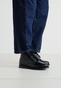 Botas de cuero negro con cordones, punta redonda y suela de goma, combinadas con pantalones de mezclilla oscuros y acampanados. Visibilidad de los cordones y acabado brillante.
